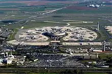 Farbfotografie in der Vogelperspektive von einem Flughafen mit runder Abfertigungshalle und rundem Außenbereich auf dem Flugzeuge sind. Im Vordergrund sind ein großer Parkplatz und eine Zugstrecke. Im Hintergrund sind Landebahnen und Felder mit Industriegebäuden auf der rechten Seite.