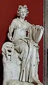 Statue von Thalia in den Vatikanischen Museen, Frontalansicht; sie sitzt auf einem Stein oder großen Baumstumpf, rechts von ihr liegt eine Theatermaske, in der linken Hand hält sie eine Handtrommel