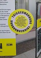 Ein rundes Schild an einer Infotafel weist darauf hin, dass die Ausstellung rassistische, antisemitische und gewaltvolle Inhalte zeigt.