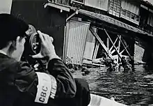 Schwarz-weiß-Bild eines Kameramanns in den Hochwasserfluten, der ein Bild von einer zerstörten Gebäudefassade macht