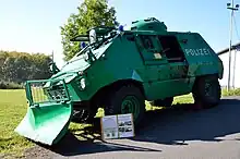 Grün lackierter, mit Räumschild, Blaulicht und Schriftzug POLIZEI versehener gepanzerter Wagen von demselben Typ wie auf dem Bild weiter oben, aufgenommen von links vorne. Die in der Fahrzeugmitte befindliche seitliche Schwenktür ist offen. Das Fahrzeug ist ein Ausstellungsstück; vor ihm ist einer Tafel mit Erläuterungen montiert. Zu erkennen sind zwei Überschriften: "Thyssen Rheinstahl UR 416 Geschützter Polizei-Sonderwagen" und "Thyssen Rheinstahl UR 416 im weltweiten Einsatz".