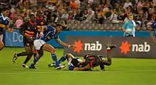Drei dunkelhäutige Rugby-Spieler laufen zu zwei anderen, die im Spielverlauf zu Boden gegangen sind.