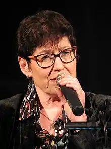 Uschi Brüning ist ein Mikrofon halten au einer Farbfotografie im Hochformat abgebildet. Sie hat kurzes braunes Haar und trägt eine Brille.