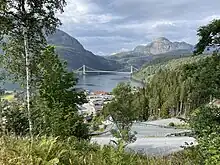Im Vordergrund ein Dorf im Grünen, im Hintergrund eine den Fjord überspannende Hängebrücke.