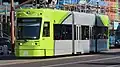 Fahrende Straßenbahn in Großaufnahme. Grau lackiertr Fahrzeugseiten, Köpfe und Teile der oberen Seiten in dynamischer, limettengrüner Signalfarbe gestaltet. Vorne als Logo als weißes, stilisiertes M.