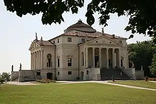 Seitliche Farbfotografie eines dreigeschossigen Gebäudes mit zwei Säuleneingängen und Giebeldächern an beiden Seiten, auf denen Figuren stehen. Zwei Außentreppen mit Figuren führen zu den Türen. Auf dem Dach ist ein flacher Kuppelaufbau. Der Vorplatz besteht aus Rasen mit Beeten und im oberen Bildrand ragen Äste.