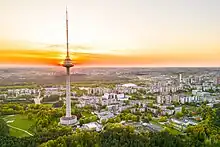 Farbfotografie von einem hohen Fernsehturm mit Sonnenuntergang. Vorne sind Grünanlagen und im Hintergrund viele Häuser.