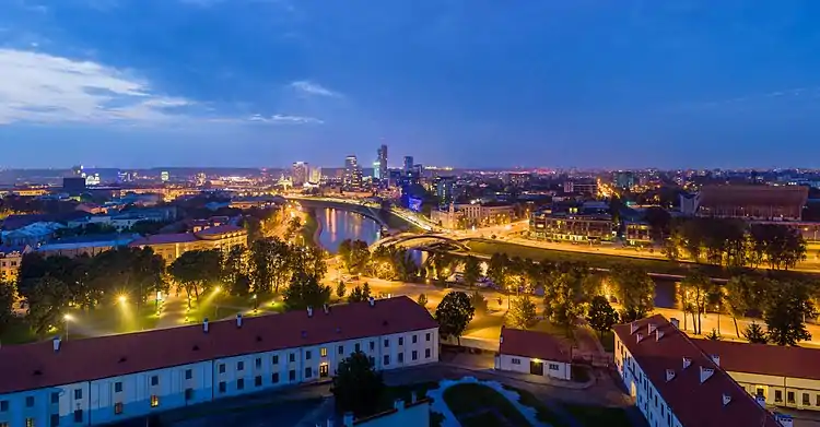 Nachtaufnahme der Stadt mit beleuchteten Straßen und Hochhäusern. Im Vordergrund sind alte Reihenhäuser und Alleen. Von rechts führt ein Fluss bis zur Bildmitte.