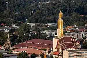 Die riesige goldfarbene Statue überragt alle Gebäude. Im Hintergrund ist bewaldetes Gebiet zu sehen.