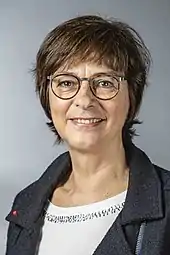 Annette Watermann-Krass hat kurze dunkelbraune Haare im Pagenkopfschnitt mit Pony und trägt eine Brille, dazu ein weißes T-Shirt mit rundem Ausschnitt zur grau-blauen grobgewebten Wolljacke.