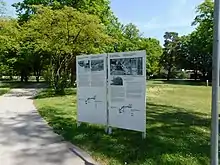 Das Foto zeigt 2 Texttafeln auf dem „Weg des Erinnerns“ in Dachau, die Tafeln stehen in der grünen Wiese, ein Weg führt daran vorbei