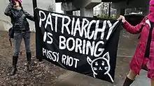 Zwei Personen halten ein schwarzes Transparent mit weißer Aufschrift: Patriarchy is boring Pussy Riot.