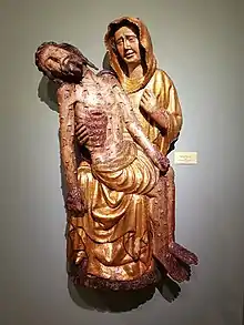 Hölzerne Darstellung einer Figur mit goldener Kleidung; sie trägt den blutüberströmten Leichnam Christis im Schoß