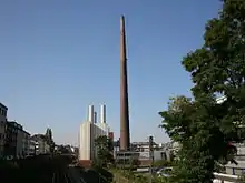 Blick über eine Eisenbahnstrecke auf einen teilweise metallverkleideten Gebäudekomplex in städtischer Umgebung. Das Foto wird in der Mitte von dem riesigen Backsteinschornstein dominiert. Links daneben ein deutlich niedrigerer, kastenförmiger, fensterloser Baublock mit heller Verkleidung und mehreren niedrigen, metallenen Schornsteinen über dem Dach.