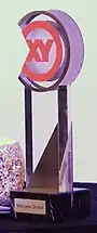 Etwa 20 Zentimeter hoher transparenter Pokal mit dem runden orangefarbenen XY-Logo oben und einem schwarzen Sockel mit messingfarbenen Plättchen mit dem eingravierten Namen „Manuela Droste“