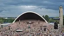 Farbfotografie in der Obersicht von einer riesigen Menschenmenge, die eine Open Air Bühne bis zur Konzertmuschel ausfüllt. Mehrere Lautsprecheranlagen sind verteilt und rechts steht ein grauer, langer Turm. An beiden seitlichen Mauern stehen aufgereiht blau-schwarz-weiße Nationalflaggen. Im linken Hintergrund ist der Hafen mit Schiff zu sehen.
