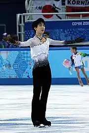 Hanyu steht mit ausgebreiteten Armen, zu einer Verbeugung ansetzend, in der Mitte der Eisfläche. Sein Kostüm besteht aus einem weißen, mit buntem Strass besetzen Hemd, einer schwarzen Hose und schwarzen Handschuhen. Im Hintergrund  ein Blumenkind vor die Bande im Design der Olympischen Spiele in Sotschi, dahinter vor der Publikumstribüne ein unterstützendes Transparent.