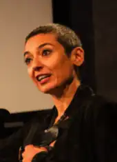  Zainab Salbi, kurze schwarz-grau-melierte Haare, trägt eine schwarze Jacke, sie steht und spricht stehend in ein Mikrophon