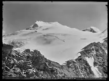 Schwarz-Weiß Fotografie eines stark vergletscherten Berggipfels. Am unteren Bildrand zieht sich ein Felsband durch das Bild, etwas rechts von der Mitte sieht man eine Berghütte.