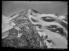Schwarz-Weiß Foto eines Schnee-, bzw. gletscherbeckten Berggipfels. Im Vordergrund leicht unscharf eine Berghütte auf felsigem Grund.