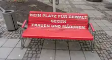 Das Foto zeigt eine Sitzbank, die rot bemalt ist und auf der Lehne in Versalien „KEIN PLATZ FÜR GEWALT GEGEN FRAUEN UND MÄDCHEN“ anzeigt