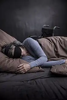 Eine an ME/CFS erkrankte Person liegt auf der Seite in einem Bett. Sie ist mit einer Decke zugedeckt. Die Person trägt einen Gehörschutz und eine Schlafmaske. Im Hintergrund steht ein Rollstuhl.